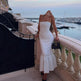 Woman in a white polka dot dress standing on a balcony with a waterfront view.