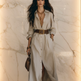 A woman models a long-sleeve linen shirt dress in a light beige color, styled with a brown belt, gold jewelry, and brown flat shoes against a plain white background.