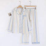 A two-piece cotton pajama set consisting of a long-sleeve button-up shirt and matching pants, hanging on wooden hangers against a plain white background. The pajamas feature a textured gauze-like fabric with vertical stripes in cream, light blue, and white, accented with light blue piping.