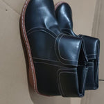 A pair of black leather ankle boots with white contrast stitching and brown soles, shown on a plain cardboard background.