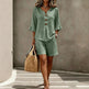 Woman wearing a green outfit with shorts and a top, holding a straw bag against a neutral background.