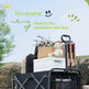 A black folding camping car cart loaded with a cooler, basket of flowers, teddy bear, and folded chairs, set outdoors on a rocky path with promotional text at the top.
