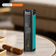 A slim rectangular cigar lighter standing upright on a round marble table, with a cigar resting nearby. The lighter has a black body with a bright teal side panel and a small clear window near the bottom.