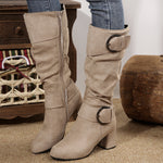 Beige knee-high boots with buckle details worn by a person, with a wooden stool and basket in the background.