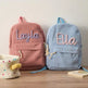 Two personalized corduroy backpacks, one dusty pink with a lavender embroidered 'Layla' and one light blue with a peach embroidered 'Ella', displayed on a wooden surface next to a polka-dot fabric basket.