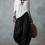 A woman modeling an off-the-shoulder white top and loose black harem-style pants, accessorized with layered necklaces, large hoop earrings, bracelets, and a woven handbag, against a textured gray studio backdrop.