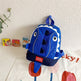 Blue shark-shaped backpack on a white wall with children's books and a plant in the background.