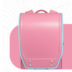 A pink school backpack with a structured flap front, light blue piping trim, and silver metal rivets, shown against a white background with a pink graphic shape behind it.