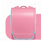 A pink school backpack with a structured flap front, light blue piping trim, and silver metal rivets, shown against a white background with a pink graphic shape behind it.