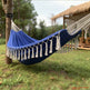A blue hammock with white tassels hanging between two trees in a grassy outdoor patio setting with a thatched-roof structure in the background.