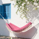 A pink fabric hammock with cream tassels hangs outdoors against a white wall with blue shutters and white flowering vines.