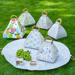 Five reusable picnic bags with floral and fruit prints are arranged on a round picnic blanket on green grass, alongside grapes, a small radio, and a sandwich. The bags have light wood handles and are shown in a real outdoor setting, making the subject type Lifestyle.