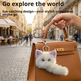 A tan leather handbag with gold hardware is carried by a person's hand on a cobblestone street. A white fluffy cat plush keychain with purple ears and blue eyes hangs from the bag's ring.