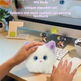 A white plush cat toy with purple ears and blue eyes is being held by two hands at a desk, with promotional text overlaid at the top about touch sensing and feedback.