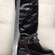 Black knee-high boot with brown belt and decorative elements, held up against a white background with a ruler for scale.