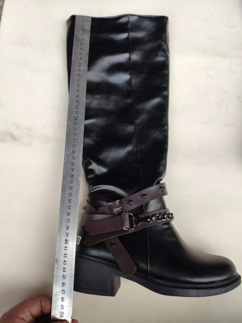 Black knee-high boot with brown belt and decorative elements, held up against a white background with a ruler for scale.