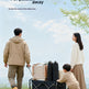 A family of three outdoors on a grassy field under a blue sky, pushing a black folding utility wagon loaded with gear. The father wears a beige jacket and pants, the mother wears a cream sweater and brown skirt, and a young child wears a beige outfit.
