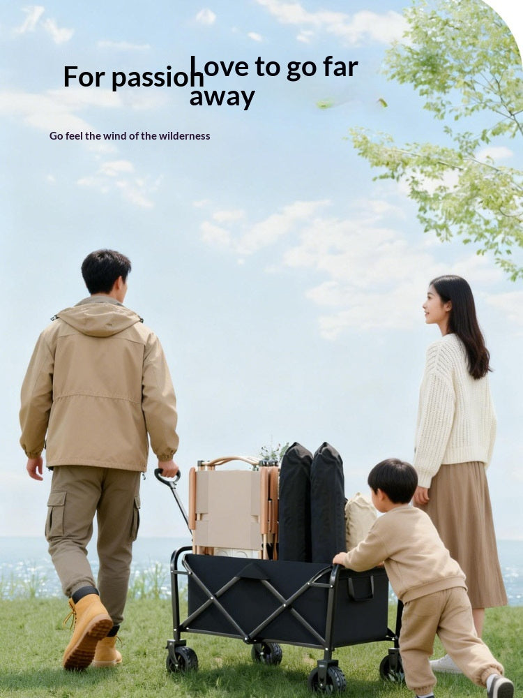A family of three outdoors on a grassy field under a blue sky, pushing a black folding utility wagon loaded with gear. The father wears a beige jacket and pants, the mother wears a cream sweater and brown skirt, and a young child wears a beige outfit.