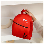 Red backpack with a cartoon face design held by a person on a white surface.