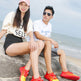 A man and a woman sitting on a large rock by the ocean, wearing bright red and yellow athletic shoes. The woman wears a cream graphic t-shirt, black shorts, and a pink cap, while the man wears a white t-shirt, sunglasses, and gray shorts.