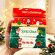 A hand holds a stack of four Christmas-themed rectangular boxes or packages decorated with festive illustrations and text including 'Merry Christmas,' 'Santa Claus,' and 'Ho Ho Ho.' The background is softly blurred with warm lights and holiday greenery.