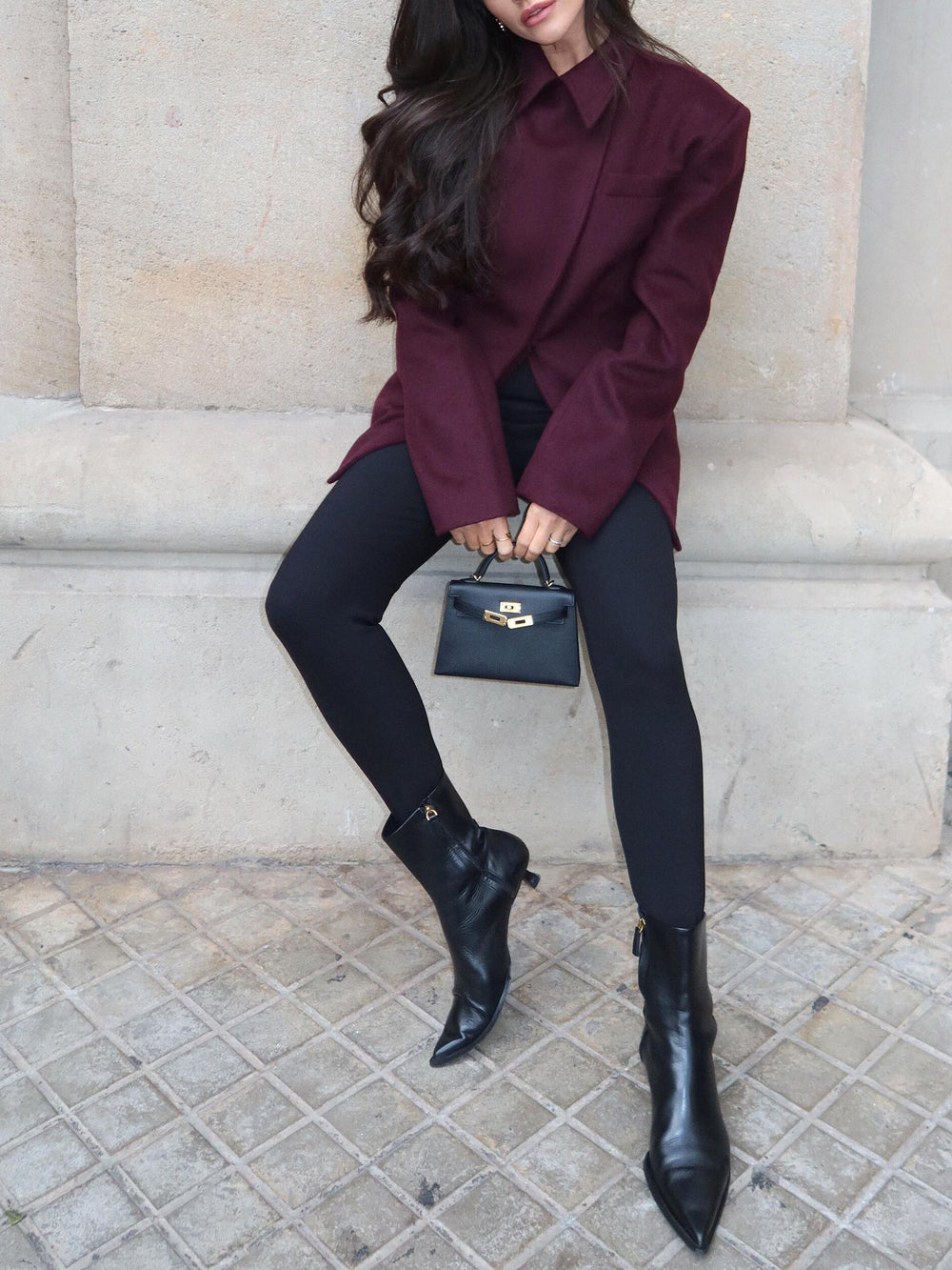 Woman wearing a burgundy coat, black leggings, and black boots sitting on a stone bench.