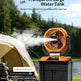 A portable ultrasonic mist fan and water tank set shown outdoors near a tent and lake, with mist spraying from the fan. The product has a black body with orange accents and a transparent tank.