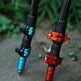Close-up of two carbon fiber tripod legs with anodized metal couplers, one in blue and one in red, standing on a concrete surface with green foliage blurred in the background.