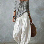 Woman wearing a gray off-shoulder top and white wide-leg pants, holding a woven bag against a gray background.