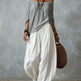 Woman wearing a gray off-shoulder top and white wide-leg pants, holding a woven bag against a gray background.
