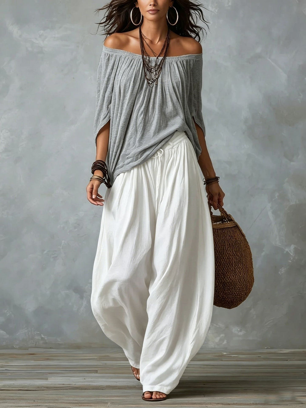 Woman wearing a gray off-shoulder top and white wide-leg pants, holding a woven bag against a gray background.