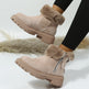 A pair of women's ankle boots in light taupe suede with fluffy faux-fur trim and a side zipper, shown being worn with black leggings. The boots feature chunky lug soles and are photographed against a light studio background with a soft beige pampas grass prop.