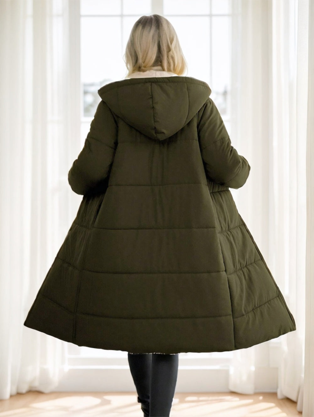 A woman viewed from behind wearing a long olive green hooded puffer coat in a bright room with white curtains and a window.