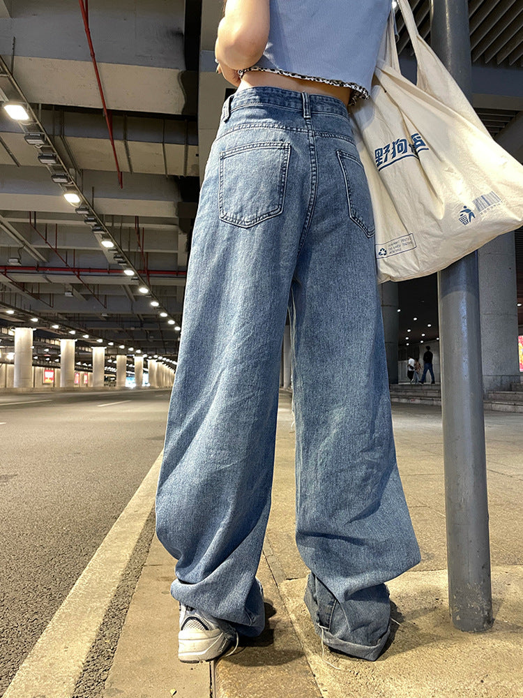 Person wearing blue jeans and a light blue shirt, standing on a sidewalk with a white bag.