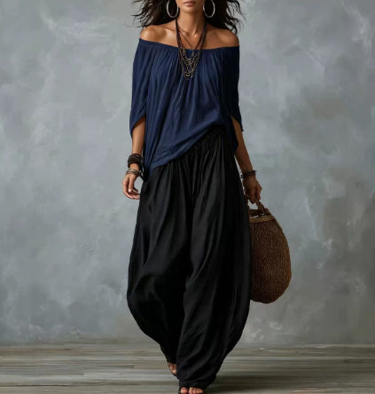 A woman models an off-the-shoulder navy blue top and black wide-leg harem pants against a textured gray studio backdrop.