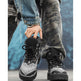 A person wearing black and white bandana-patterned lace-up boots, with one boot being held by a hand. The person is also wearing gray camouflage pants and a blue denim jacket, set against a textured blue background.