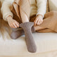 Persona con un maglione beige e pantaloni marroni seduta su un divano beige, che tiene una calza marrone.