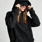 A woman wearing a black pullover hoodie with the hood pulled up over her eyes, smiling and holding the hood in place against a light gray studio background.