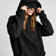 A woman wearing a black pullover hoodie with the hood pulled up over her eyes, smiling and holding the hood in place against a light gray studio background.