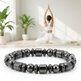 A black beaded bracelet made of polished metallic beads is shown in sharp focus in the foreground, while a woman in white yoga clothing practices in a bright, sunlit room with potted plants in the background.