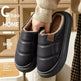 A pair of black winter slip-on slippers with plush tan lining, held by a person wearing a beige sweater. The background shows a cozy wooden interior.