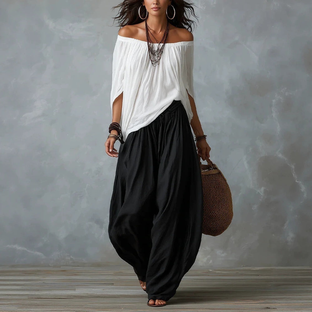 Woman wearing a white off-shoulder top and black wide-leg pants, holding a brown bag against a gray background.
