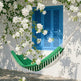 A green hammock with white fringe hangs in front of a white wall with a bright blue shutter, surrounded by white flowering vines and leaves.