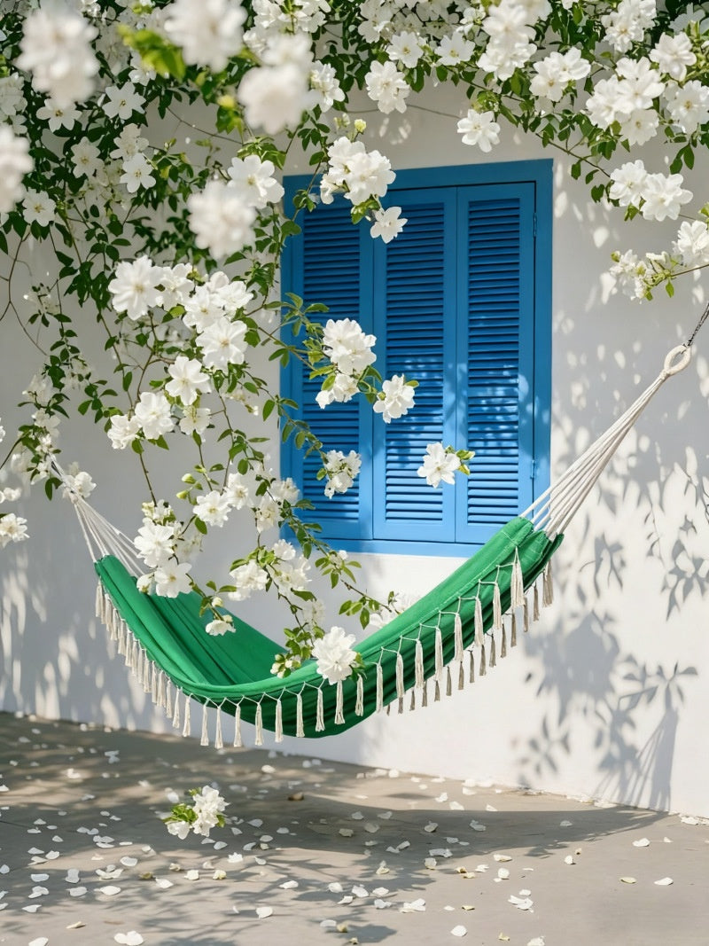 A green hammock with white fringe hangs in front of a white wall with a bright blue shutter, surrounded by white flowering vines and leaves.
