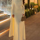 Woman in a white evening gown standing outdoors with decorative lighting and plants.