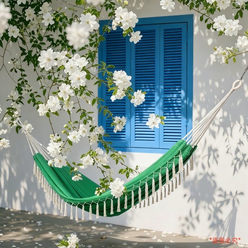 A green fabric hammock with white tassels hangs in front of a white wall and bright blue shutter window, surrounded by white flowers and leafy vines casting shadows on the wall.