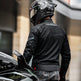 Person wearing a black motorcycle jacket and helmet standing next to a motorcycle.