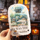 Product packaging with scenic design and text held in front of a cozy café setting