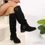 Black knee-high boots with silver buckles worn by a person sitting on a white surface.
