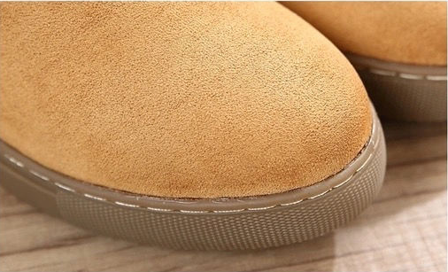 Close-up of a tan suede slipper or boot toe with a textured gray sole, resting on a light wood surface.